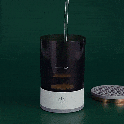 A hand pours water into a Unik Store Automatic Electric Makeup Brush Cleaner, filling it to the MAX line as the top cover rests next to the black and white device on a green surface.